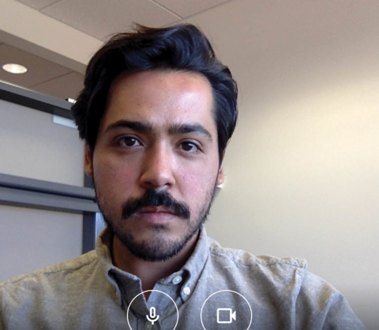A portrait of artist Sergio Serrano, which is a screenshot from a video calling app. Sergio wears a light grey shirt, with black hair and a beard and moustache, and is in a white walled room.