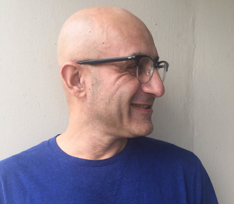 A portrait of artist Riaz Mehmood, looking to the right and smiling. Riaz has a shaved head and wears glasses and a blue shirt.