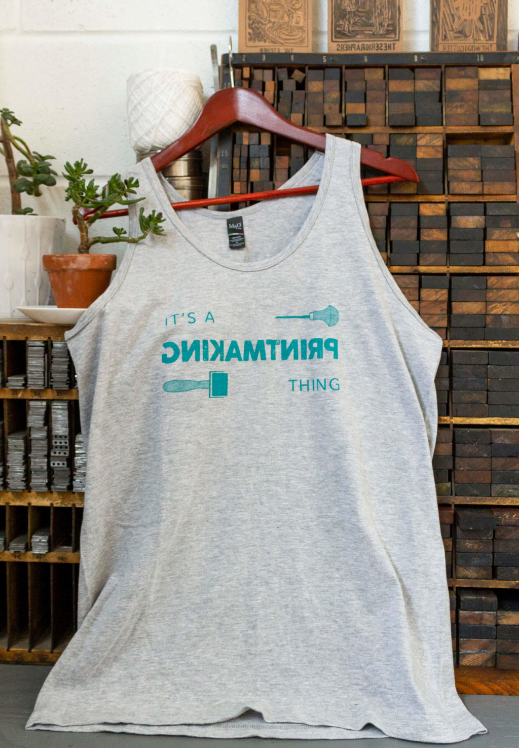 Grey tank top with words "It's a printmaking thing", with the word "Printmaking" written backwards. The text is flanked by illustrations of a lino carving tool and a brayer all printed in the colour of green emulsion.