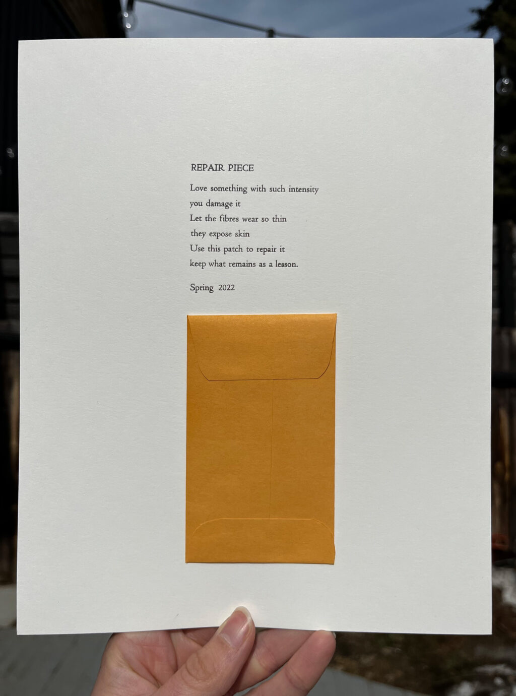 A hand holding a letterpress print, printed with a poem that reads: "REPAIR PIECE. Love Something with such intensity that you damage it. Let the fibres wear so thin they expose skin. Use this patch to repair it. keep what remains a lesson. Spring 2022" The poem is followed by a small manila envelope glued onto the paper.