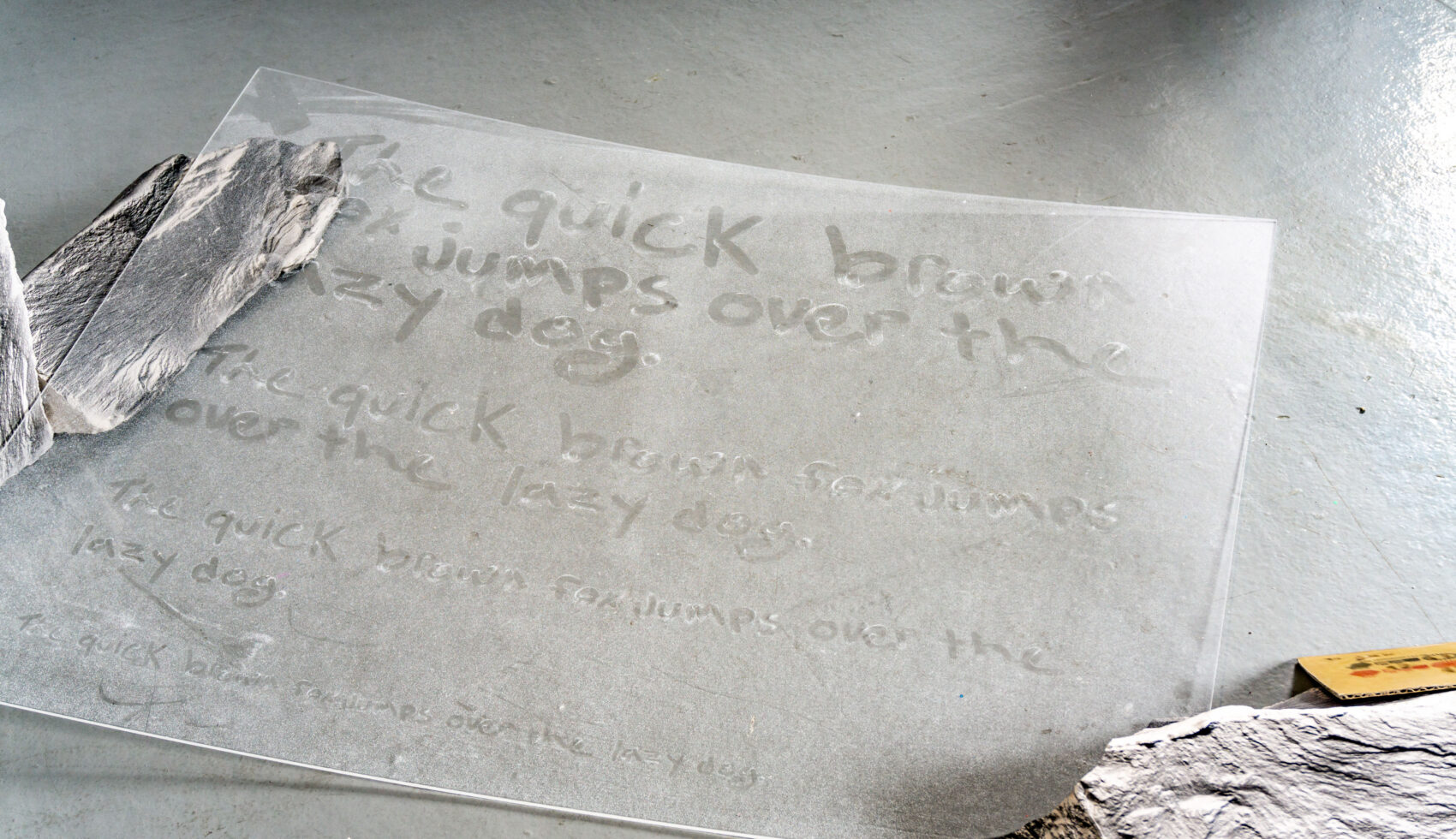 Artwork by Alex Linfield. A sheet of plexiglass on a floor supported by ceramic castings of rocks. The plexiglass appears to have text drawn into the dust with a finger, which is in fact a screen printed layer.