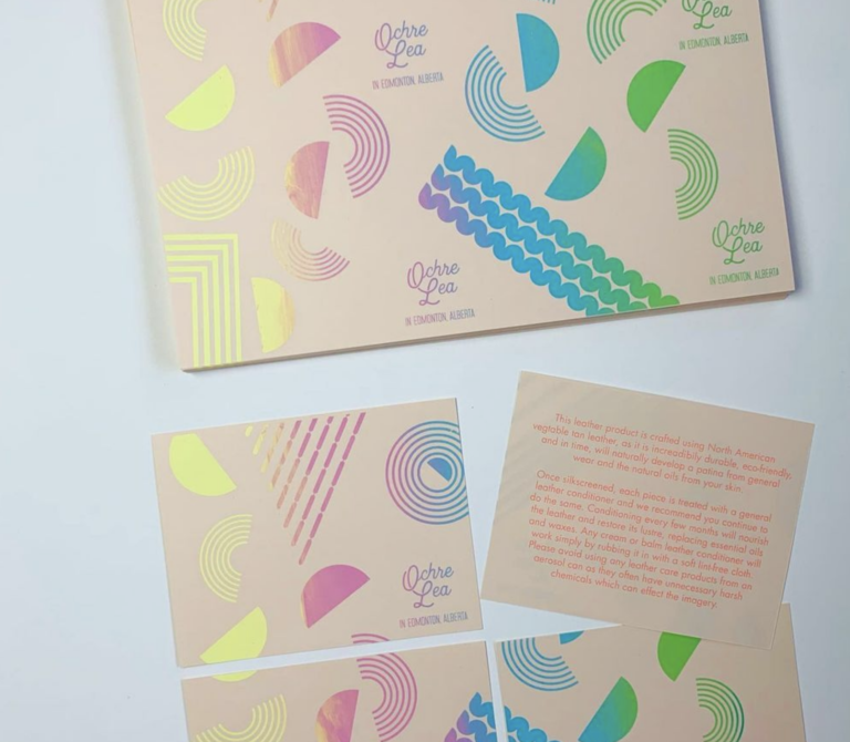 A photo of tan coloured cards lying on a white surface, with a geometric pattern of semi-circles and wiggles printed in a rainbow gradient. On the cards there is the text ‘Ochre Lea along In Edmonton, Alberta’.