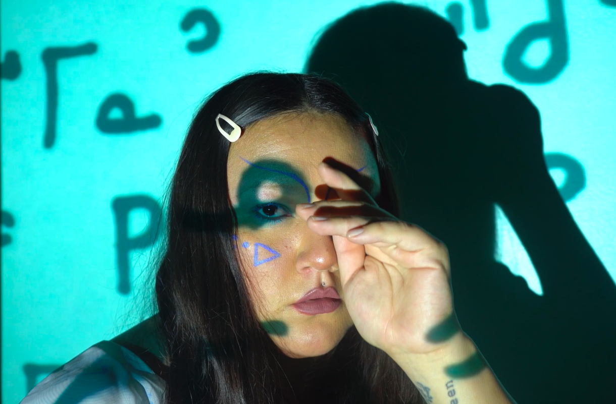 A still from the performance "Cahkipêhikan". An Indigenous woman stares at the camera wearing two pink clips in her hair. She has blue Nehiyaw (Cree) syllabics drawn on her face. Projected on her face and the wall is Nehiyaw Syllabics meaning: Mosum and Kokum.