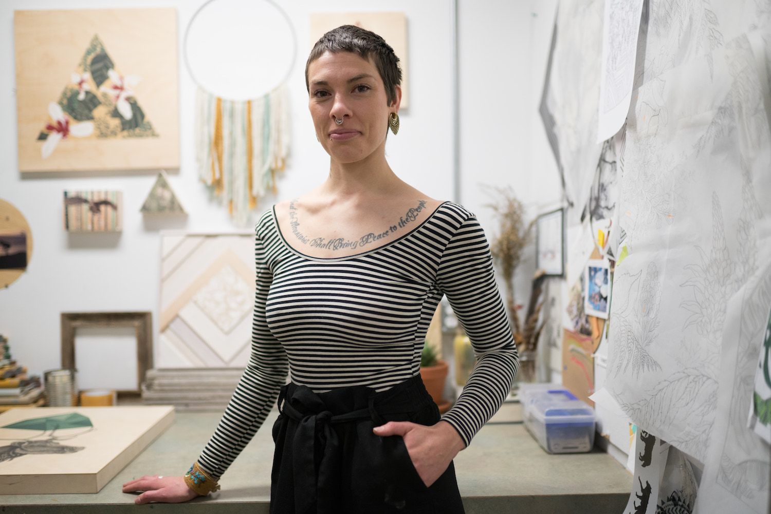 A portrait of artist Caitlin Bodewitz standing in her studio, wearing a striped shirt and black pants, with close cropped black hair.