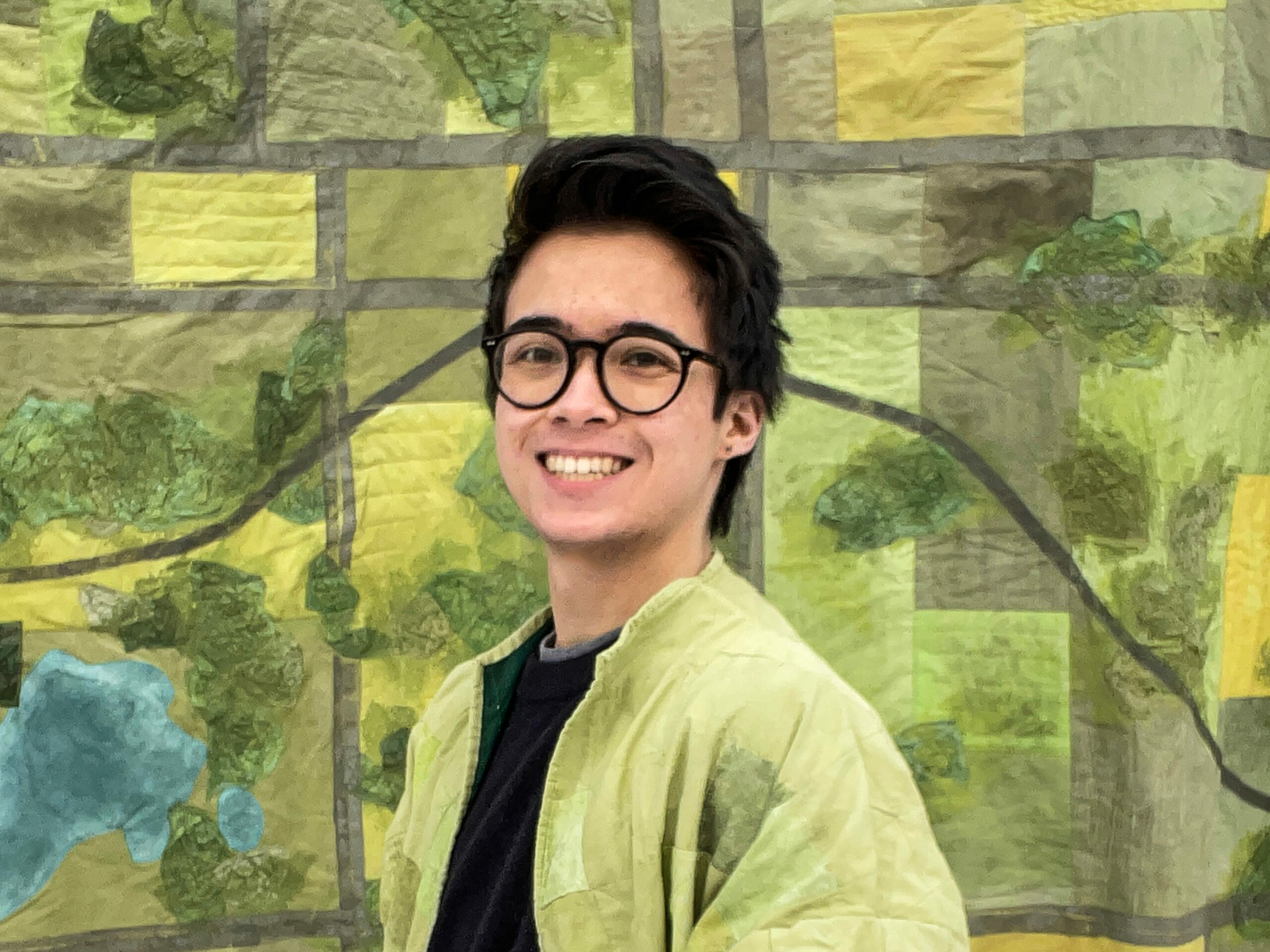a portrait of Devin smiling at the camera and posing with their hands in their pockets. They are wearing a light green jacket and black shirt and pants, and has round black glasses and short black hair. In the background is a quilted fabric piece with various green and grey textures.
