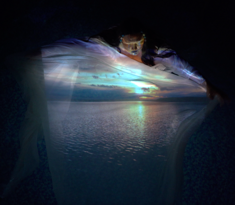A still from the performance Grieving with the Land. An indigenous woman is standing in a pool of water. She has a sheer white fabric she is holding on top of the water. Projected onto the sheer fabric, water and her body is an image of the water and sunset back home (Wabasca).