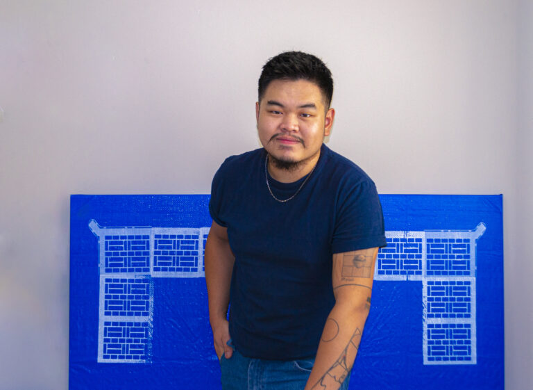 Pictured is Kev, a Chinese-Canadian male with short and faded black hair as well as black goatee-like facial hair. He is wearing a dark blue T-shirt with a silver necklace and he is standing while leaning forward into his tattooed left arm that’s holding onto a chair (chair not pictured). Behind Kev is a previous artwork of his that consists of white Chinese design motifs printed onto the perimeters of a blue tarp, stretched onto a square canvas. 
