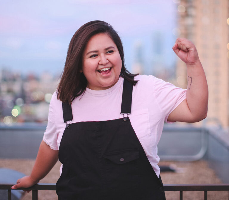 A headshot of Michelle Campos Castillo where she is standing on a rooftop, wearing a pink shirt and black overalls, sporting short dark brown hair. She is laughing and holding up her left fist.