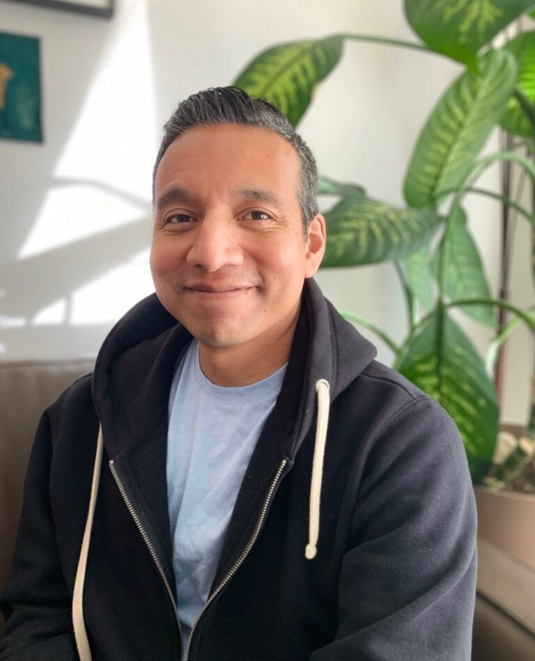 A headshot of Roger Garcia smiling at the camera, wearing a black hoodie and a blue shirt and short black hair. In the background is a living room corner with a large plant.