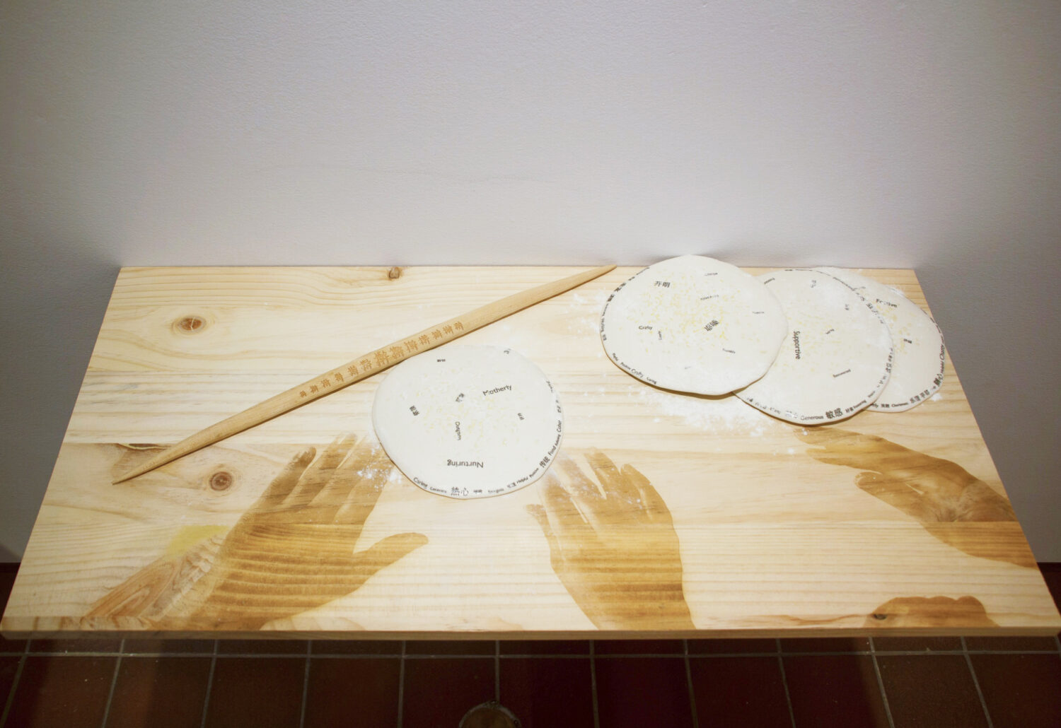 An art installation featuring a wood board with images of hands engraved on it, with flat round pieces of dough laid out on the surface.