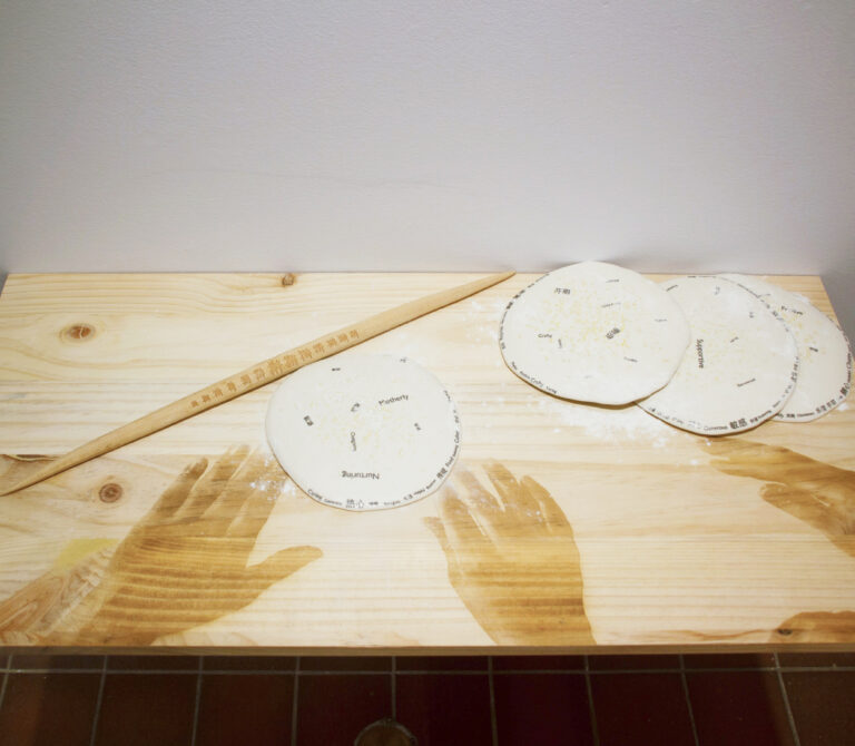 An art installation featuring a wood board with images of hands engraved on it, with flat round pieces of dough laid out on the surface.