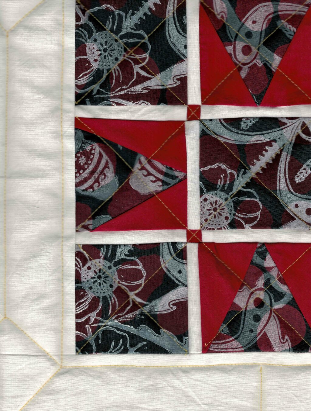 Cropped view of a quilt with squares of printed fabric and red triangles forming a square star. The squares feature a deep red bAckground and dark blue floral patterns reminiscent of Ukrainian traditional art.