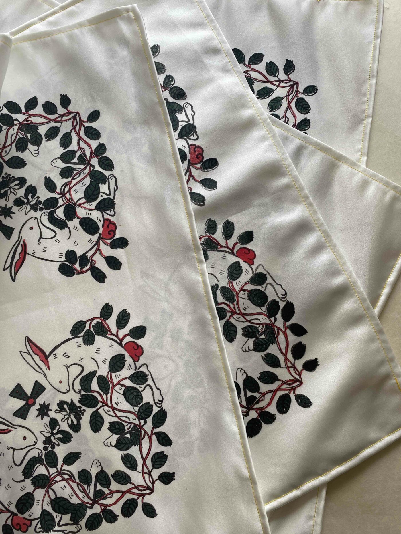A layered set of 4 hemmed pieces of satin with the bottom 3 partially obscured. They are screenprinted with a rabbit and saskatoon berry motif in each corner using a black outline and red and green accents.