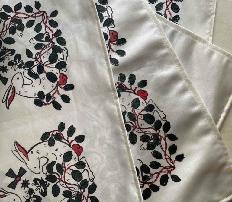 A layered set of 4 hemmed pieces of satin with the bottom 3 partially obscured. They are screenprinted with a rabbit and saskatoon berry motif in each corner using a black outline and red and green accents.