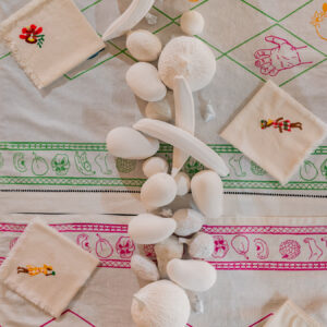 An aerial, close up view of a table with tablecloth, napkins, and white fruit made of plaster cast. The table cloth has linework drawings in green, pink and yellow with fruit, hand and feet motifs. The napkins are embroidered with figures of plantation workers.