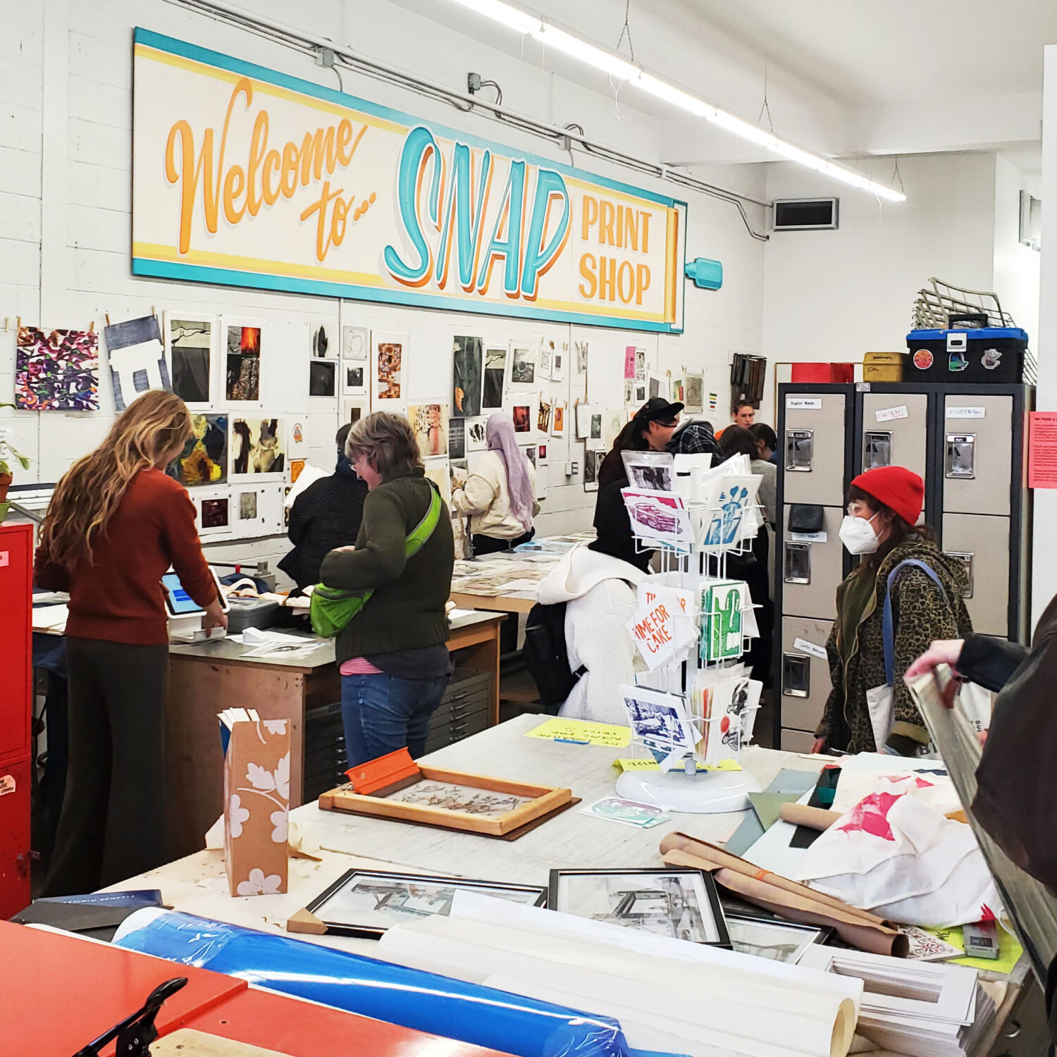 A photo of the Garage Sale taking place in the printshop, with artwork on all the walls and tables, and people browsing through them.